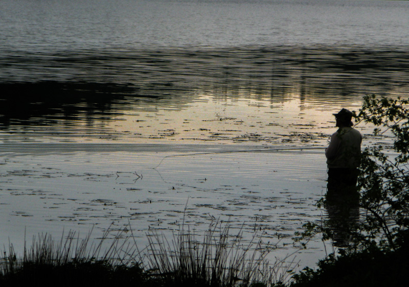 A fisherman stands waist deep in a resevoir in the late afternoon.